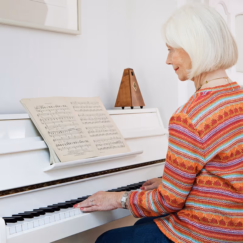 An elderly woman with white hair wearing a colorful striped sweater is playing a white piano with sheet music and a wooden metronome on top.