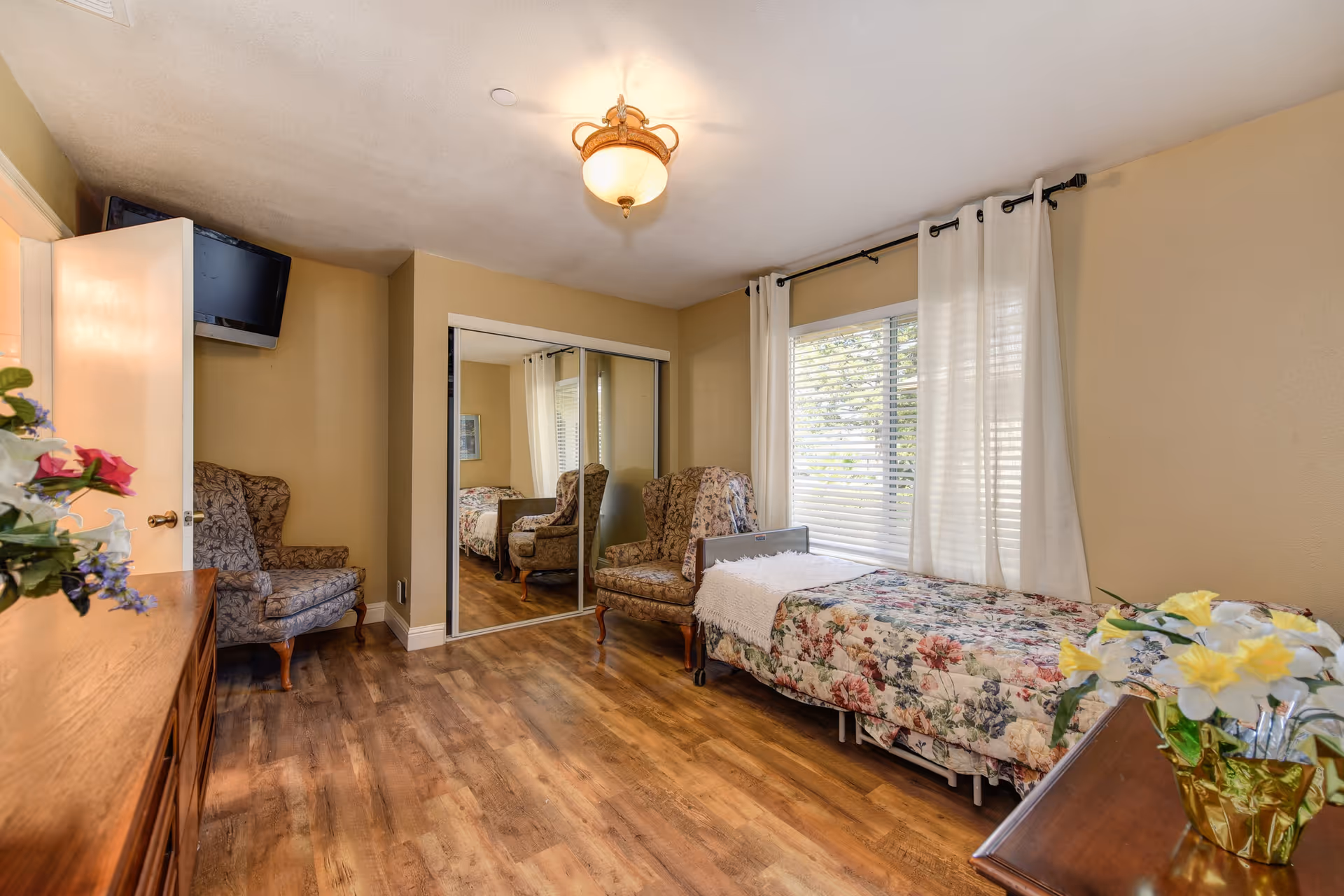 A cozy bedroom with a single bed covered in a floral bedspread, a cushioned armchair with a floral pattern, a wooden dresser with a vase of flowers, a wall-mounted TV, a window with white curtains, and a mirrored closet door reflecting part of the room.