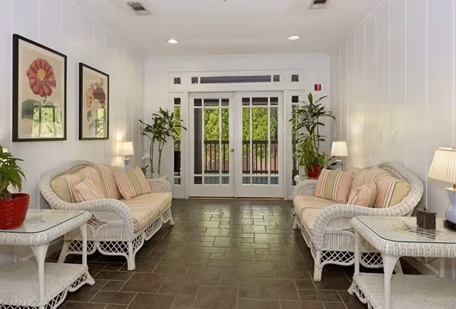 A bright and inviting living room with two white wicker sofas featuring striped cushions and pillows, two matching wicker side tables with lamps, potted plants, framed floral artwork on the walls, and glass double doors leading outside.