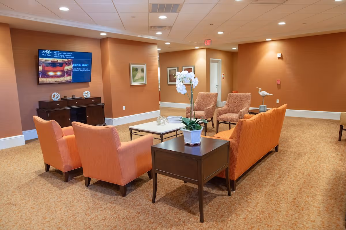 A cozy seating area in a senior living facility with orange upholstered chairs and a sofa arranged around a square coffee table. A small side table with a white orchid plant is in the foreground. The walls are painted a warm orange color, and a flat-screen TV is mounted on the wall above a dark wooden cabinet. The carpet is beige, and there are framed pictures on the walls.