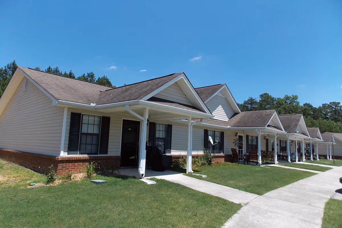 Row of single-story attached senior living units with covered front porches, a sidewalk, and a grassy lawn under a clear blue sky.