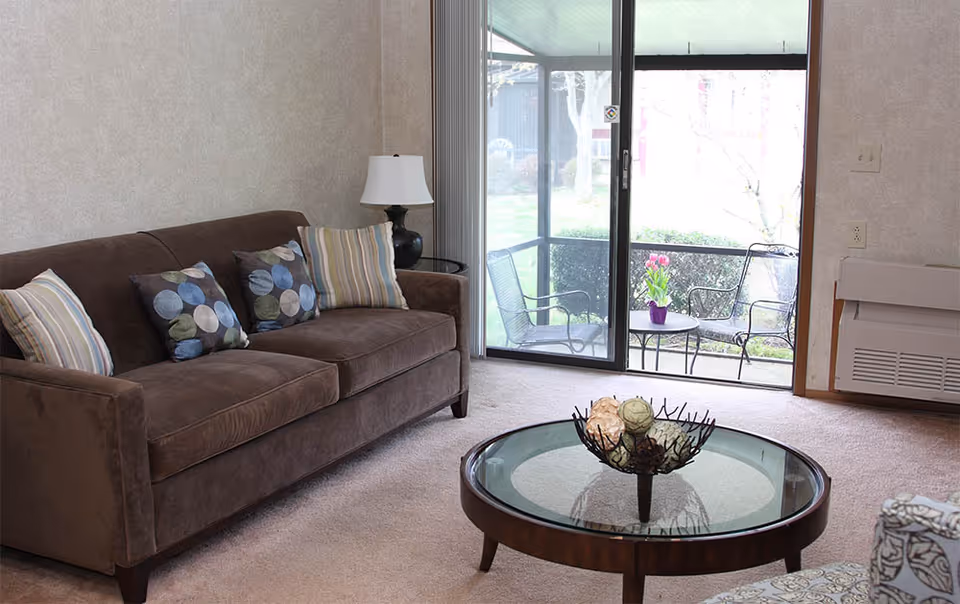 A cozy living room with a brown sofa adorned with patterned and striped pillows, a round glass coffee table with decorative items, a black table lamp on a side table, and a sliding glass door leading to an outdoor patio with metal chairs and a small table holding a purple vase with pink flowers.
