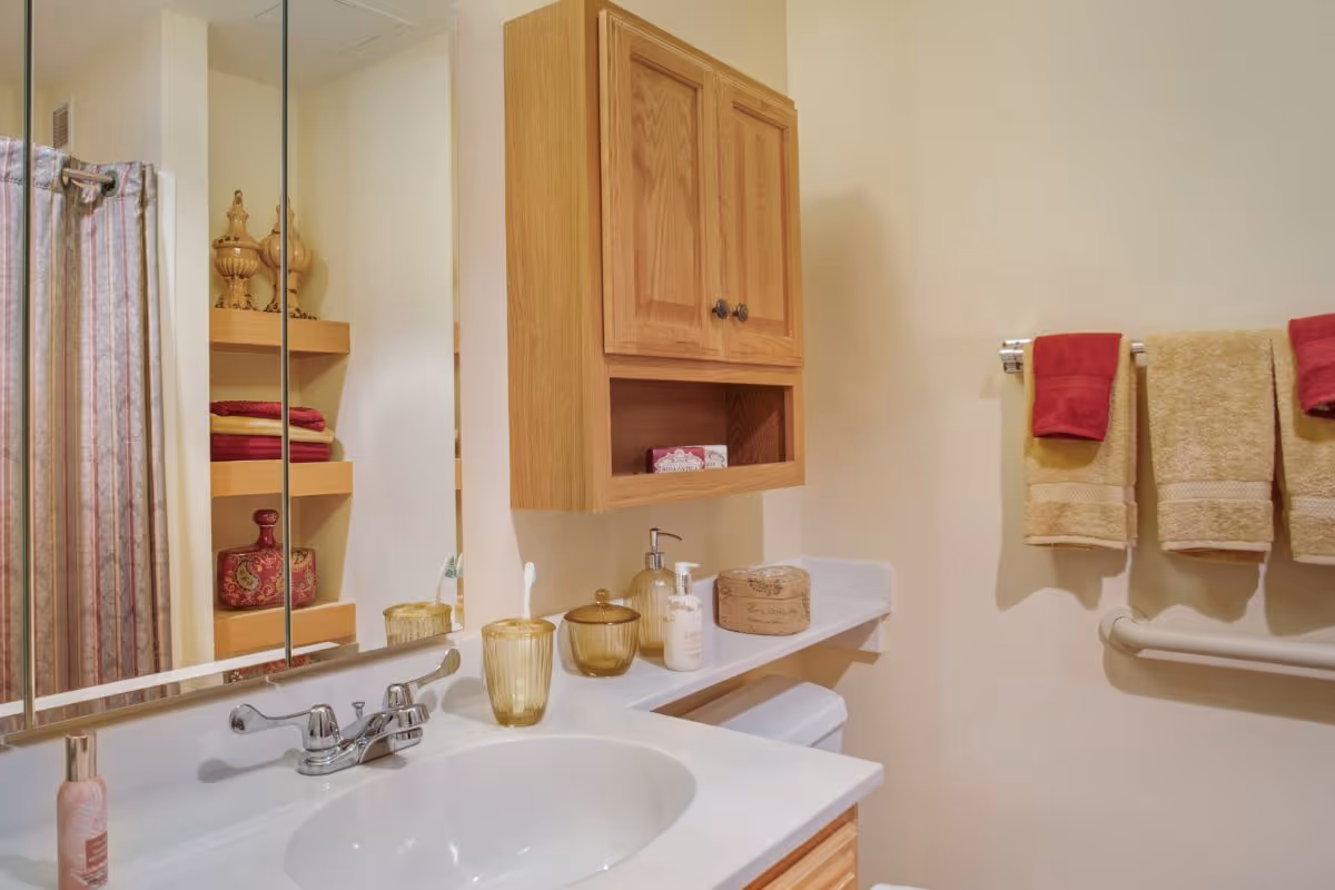 Bathroom sink and countertop with a wall-mounted wooden cabinet, mirror, toiletries, and towels hanging on a rack.