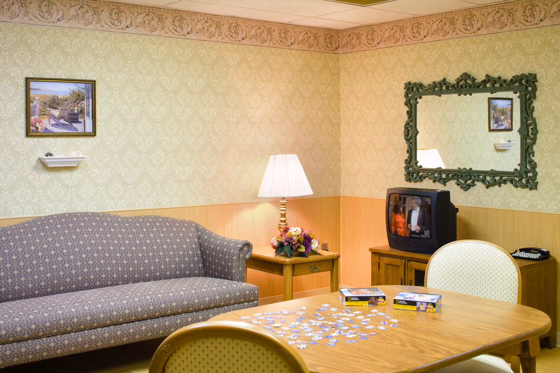 A cozy sitting area with a patterned sofa, wooden table scattered with puzzle pieces, a lamp on a side table, small TV, and a decorative mirror on wallpapered walls.