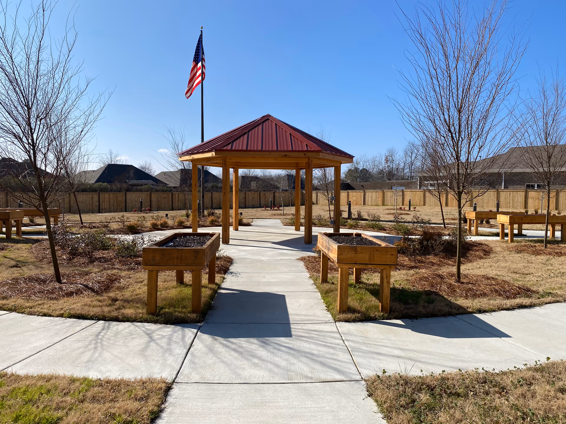 Outdoor garden area at Community Place Nursing Home featuring a paved walkway leading to a wooden gazebo with a red roof. Raised wooden planter boxes are positioned along the walkway, and an American flag is flying on a flagpole behind the gazebo. Leafless trees and a wooden fence surround the area under a clear blue sky.