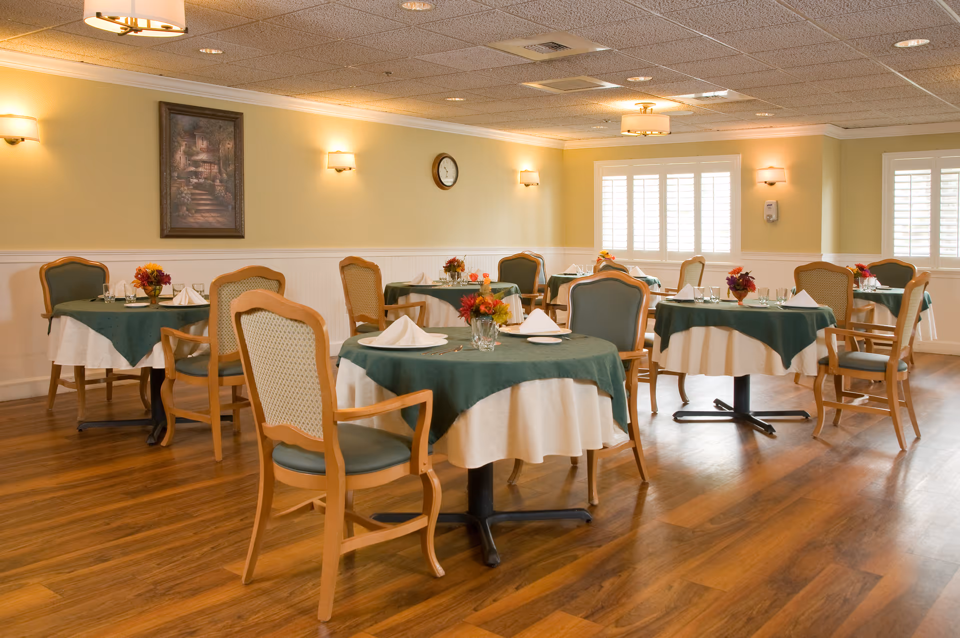 A dining room with several round tables covered with white and green tablecloths, each set with napkins, glasses, and small floral centerpieces. The room has wooden floors, beige walls with white wainscoting, wall sconces, a clock, and a framed painting. Windows with white shutters allow natural light into the space.