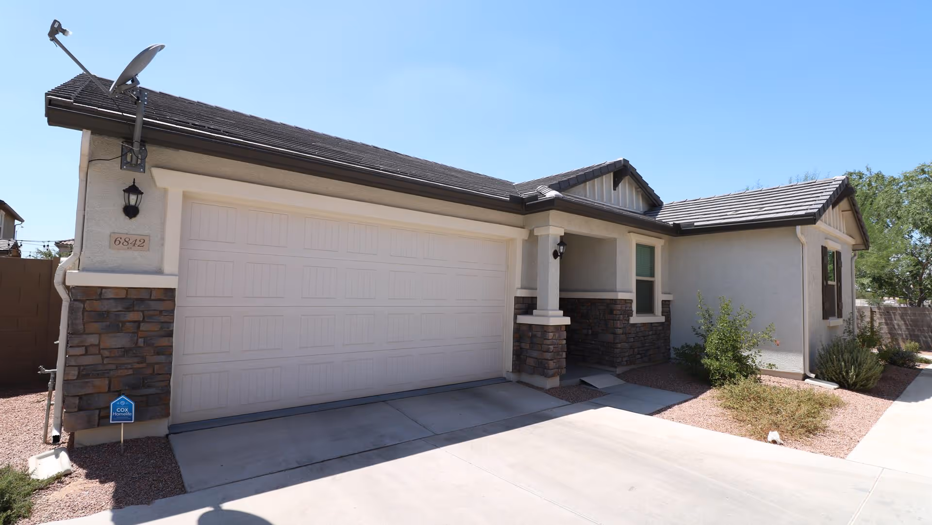 Exterior view of a single-story residential building with a two-car garage, stone accents on the walls, a small covered porch, and desert landscaping with shrubs and gravel. The sky is clear and blue.