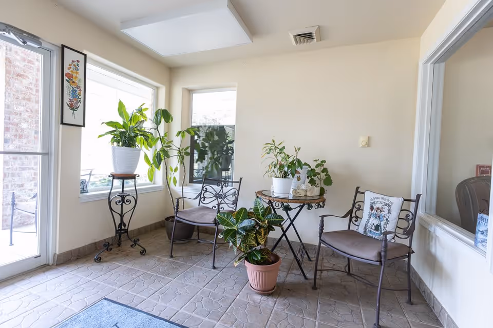 A small indoor seating area with two metal chairs with cushions, a round table with potted plants, and additional potted plants on stands near large windows letting in natural light. The floor is tiled and the walls are light-colored.