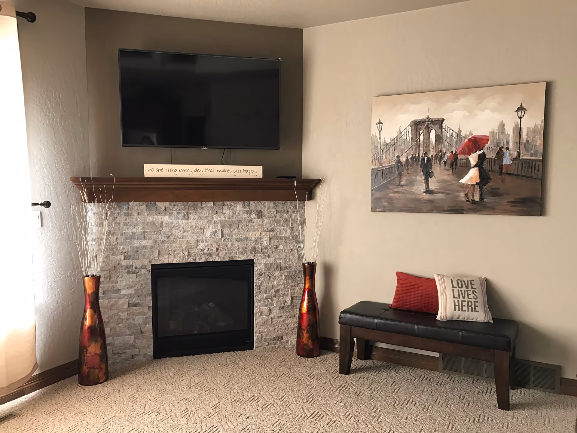 A cozy living room corner featuring a stone fireplace with a wooden mantel, a flat-screen TV mounted above it, and a decorative sign on the mantel that reads 'do one thing every day that makes you happy'. On either side of the fireplace are tall, red and orange vases with decorative branches. To the right, there is a black bench with two pillows, one red and one beige with the text 'LOVE LIVES HERE'. Above the bench hangs a painting of people walking on a bridge, with one couple holding a red umbrella.