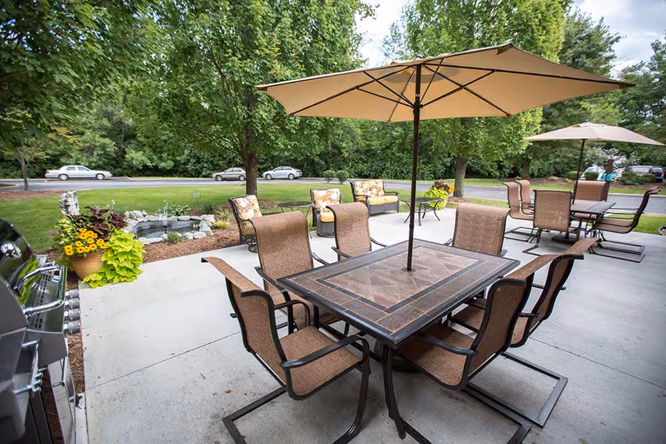 Outdoor patio area with two tables each surrounded by six chairs and large beige umbrellas. There is a small garden with flowers and a water feature, and trees and parked cars are visible in the background.