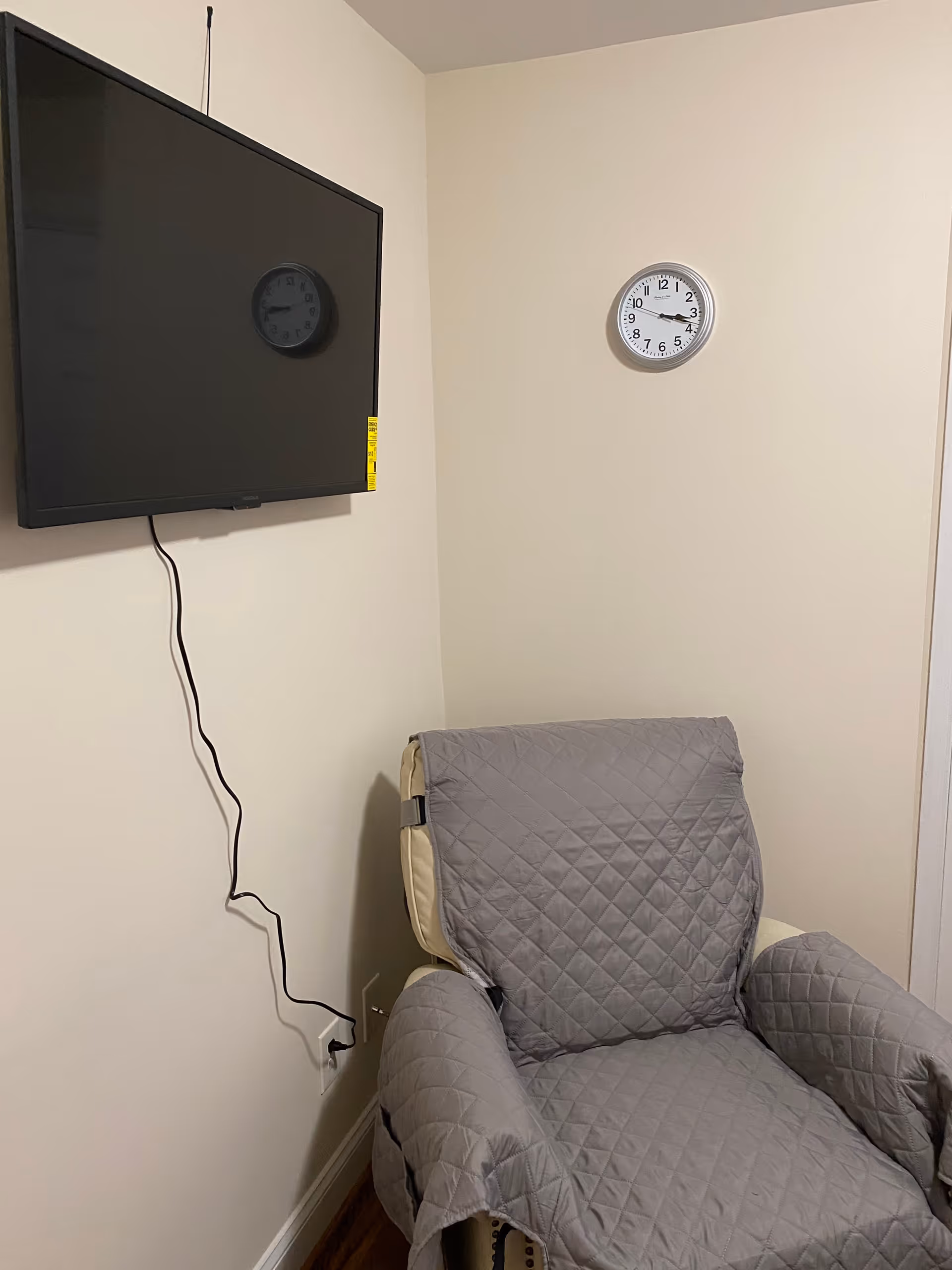 Wall-mounted TV and clock above a gray-quilted covered recliner in a small room.