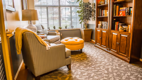 Cozy seating area with armchairs, an orange ottoman, a floor lamp, and a wooden bookcase by a large window.