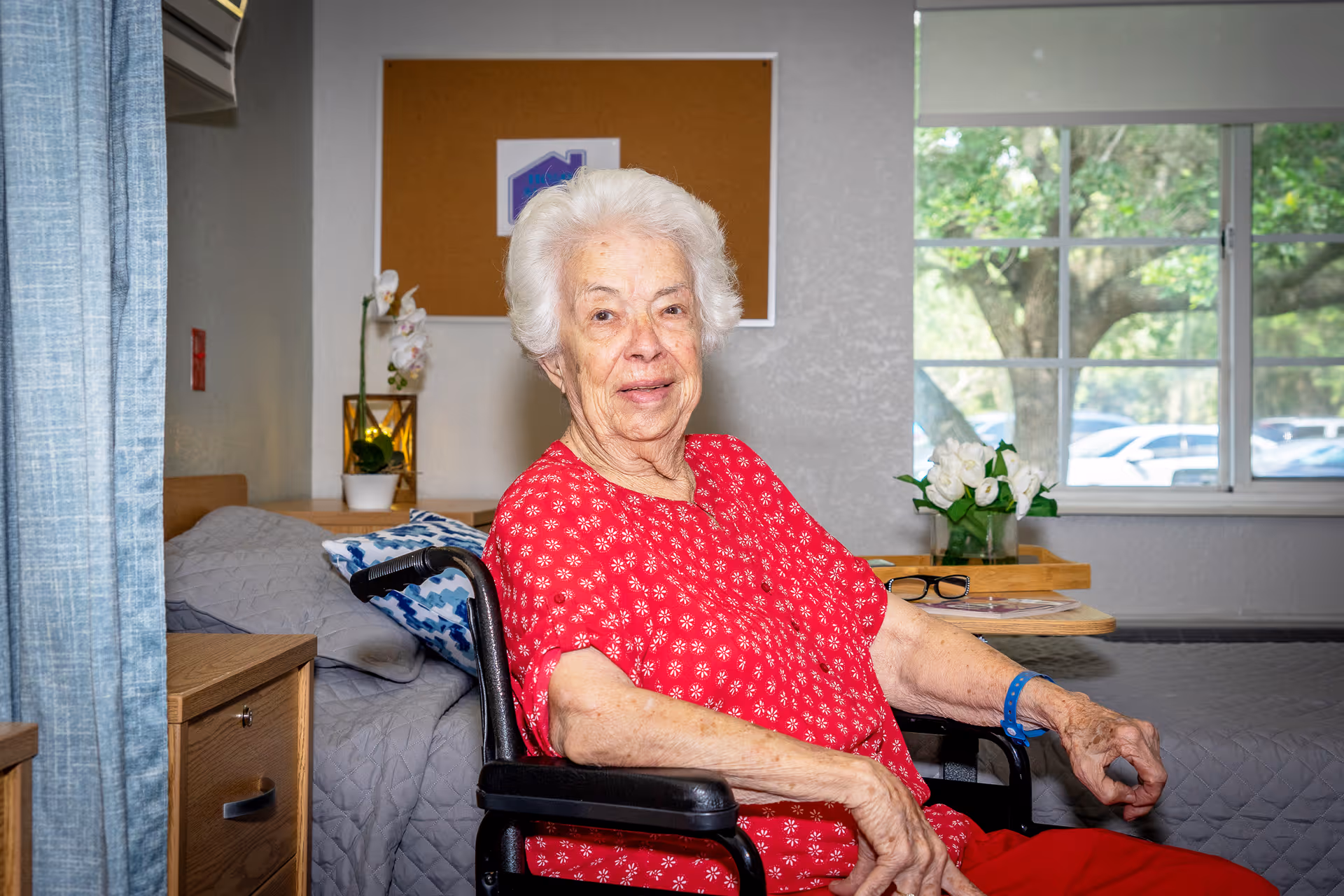 An elderly woman with white hair wearing a red patterned shirt sits in a wheelchair inside a well-lit room. Behind her is a bed with a gray quilt and a blue patterned pillow, a wooden nightstand with a potted orchid, and a window showing green trees and parked cars outside. A corkboard with a small poster is mounted on the wall.