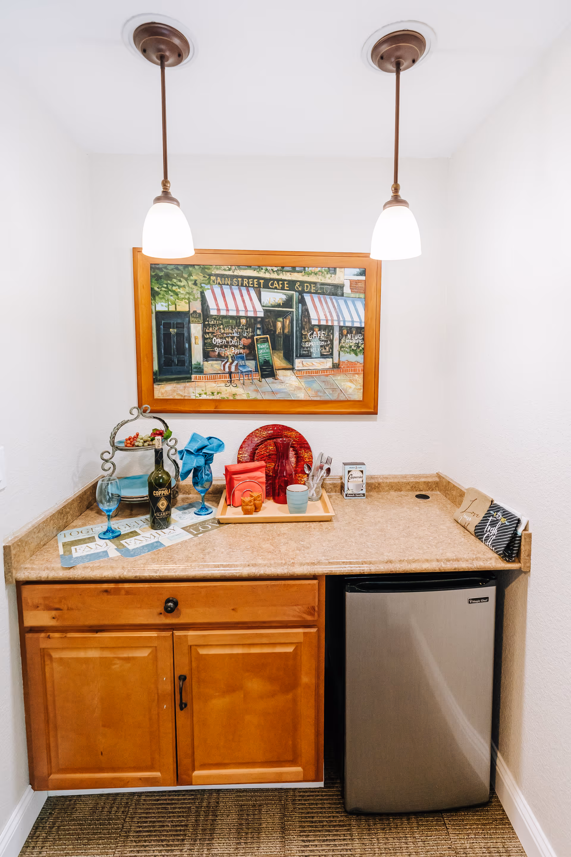 A small kitchenette area with a wooden cabinet and a mini stainless steel refrigerator. The countertop holds decorative items including a bottle of wine, two blue wine glasses, a tiered tray with grapes, a red plate, a small tray with utensils, and a framed painting of a cafe on the wall above. Two pendant lights hang from the ceiling illuminating the space.