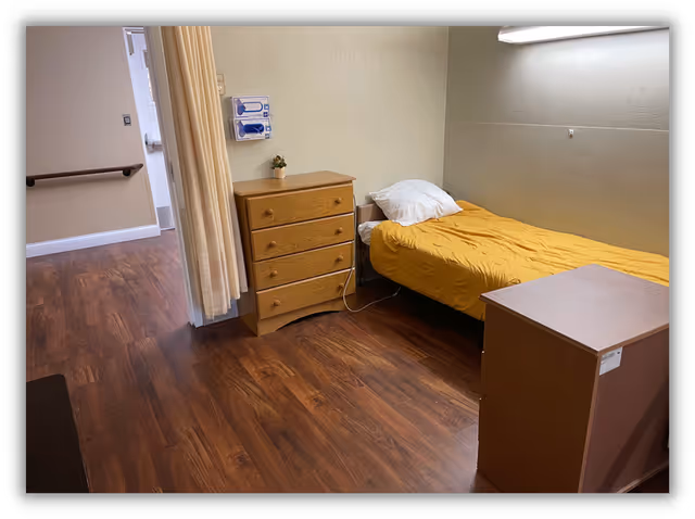 A small senior living bedroom with a single bed covered in a yellow blanket and a white pillow. Next to the bed is a wooden chest of drawers with a small plant on top. The room has wood flooring and beige walls, with a curtain partially drawn near the entrance. There is a handrail along the hallway visible outside the room.