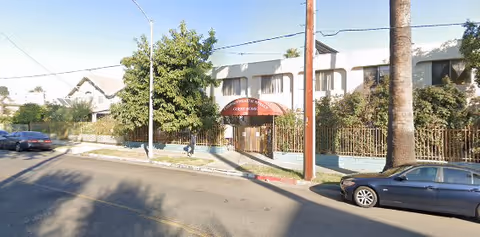 Street view of Commonwealth Royale Senior Living building with a gated entrance and a red awning displaying the facility's name. There are trees and bushes along the fence, and cars parked on the street in front of the building.