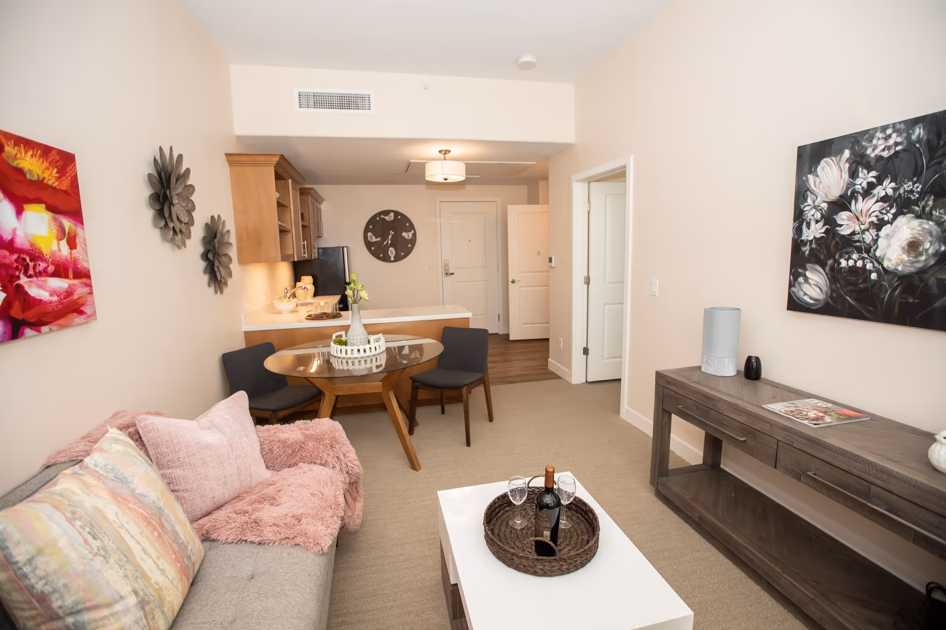 Bright furnished living room with a sofa, coffee table, round dining table, and a small kitchenette in the background.
