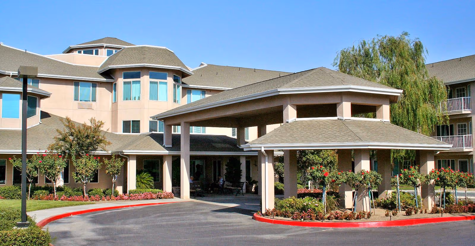 Exterior view of a senior living facility with a covered entrance driveway, landscaped bushes and flowers, and a multi-story building with large windows under a clear blue sky.