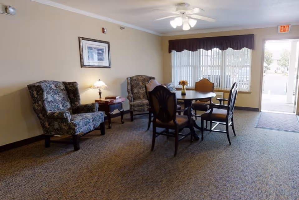 A cozy sitting area in a senior living facility with two floral upholstered armchairs, a small wooden side table with a lamp and books, and a round wooden table surrounded by four chairs. A window with vertical blinds and a valance lets in natural light, and an exit door is visible in the background.