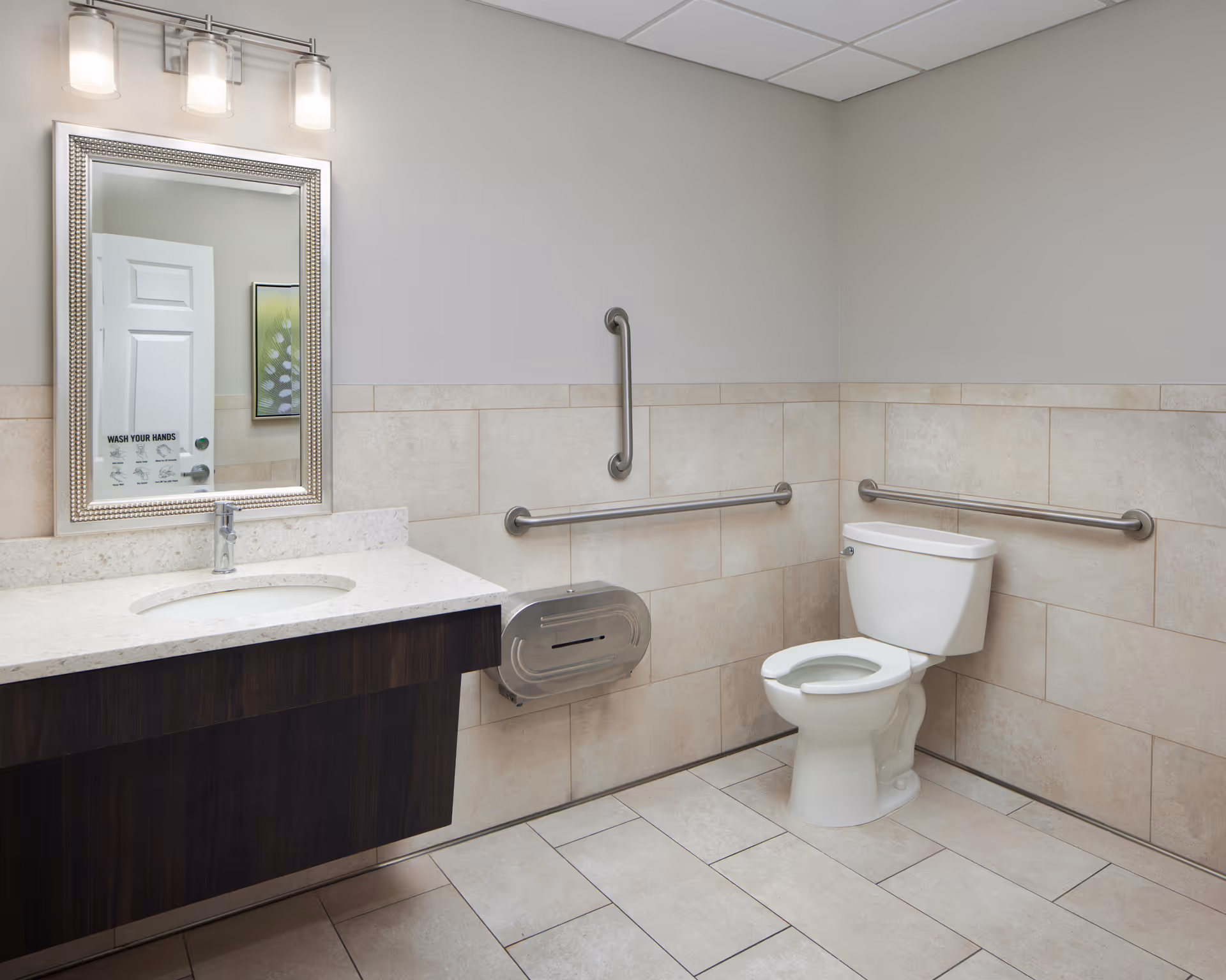 A clean, accessible bathroom with beige tiled walls and floor. The room features a white toilet with grab bars on the walls around it, a dark wood vanity with a white countertop and an oval sink, a large framed mirror above the sink, and a three-light fixture mounted above the mirror. A sign on the mirror reads 'WASH YOUR HANDS'.