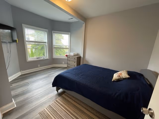 A bedroom with a bed covered in a dark blue blanket and a decorative pillow. The room has light gray walls and wood flooring with a striped rug. There are two windows with white blinds letting in natural light, a small wooden dresser in the corner, and a wall-mounted TV on the left side.