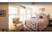 A cozy bedroom with a large bed covered in a colorful patchwork quilt, a cushioned armchair with a patterned pillow, three windows letting in natural light, a framed picture on the wall, and a bedside table with a lamp.