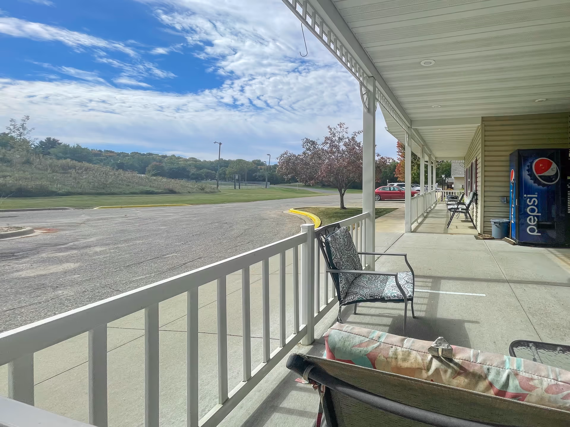 Covered outdoor patio area with several cushioned chairs and a Pepsi vending machine, overlooking a parking lot and grassy area with trees under a partly cloudy sky.