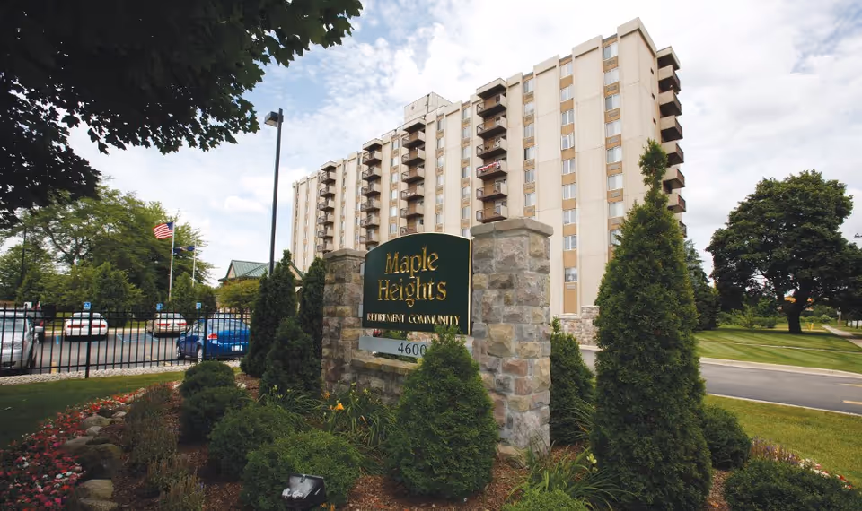 Exterior view of Maple Heights Senior Living retirement community showing a multi-story building with balconies, a landscaped garden with bushes and flowers, a stone sign with the facility name, and a parking area with cars and an American flag.