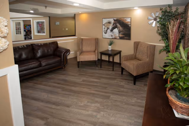 A cozy seating area in a senior living facility with a dark brown leather sofa, two beige upholstered armchairs, a small black side table with a potted plant, a framed picture of horses on the wall, and various green plants in the corner.