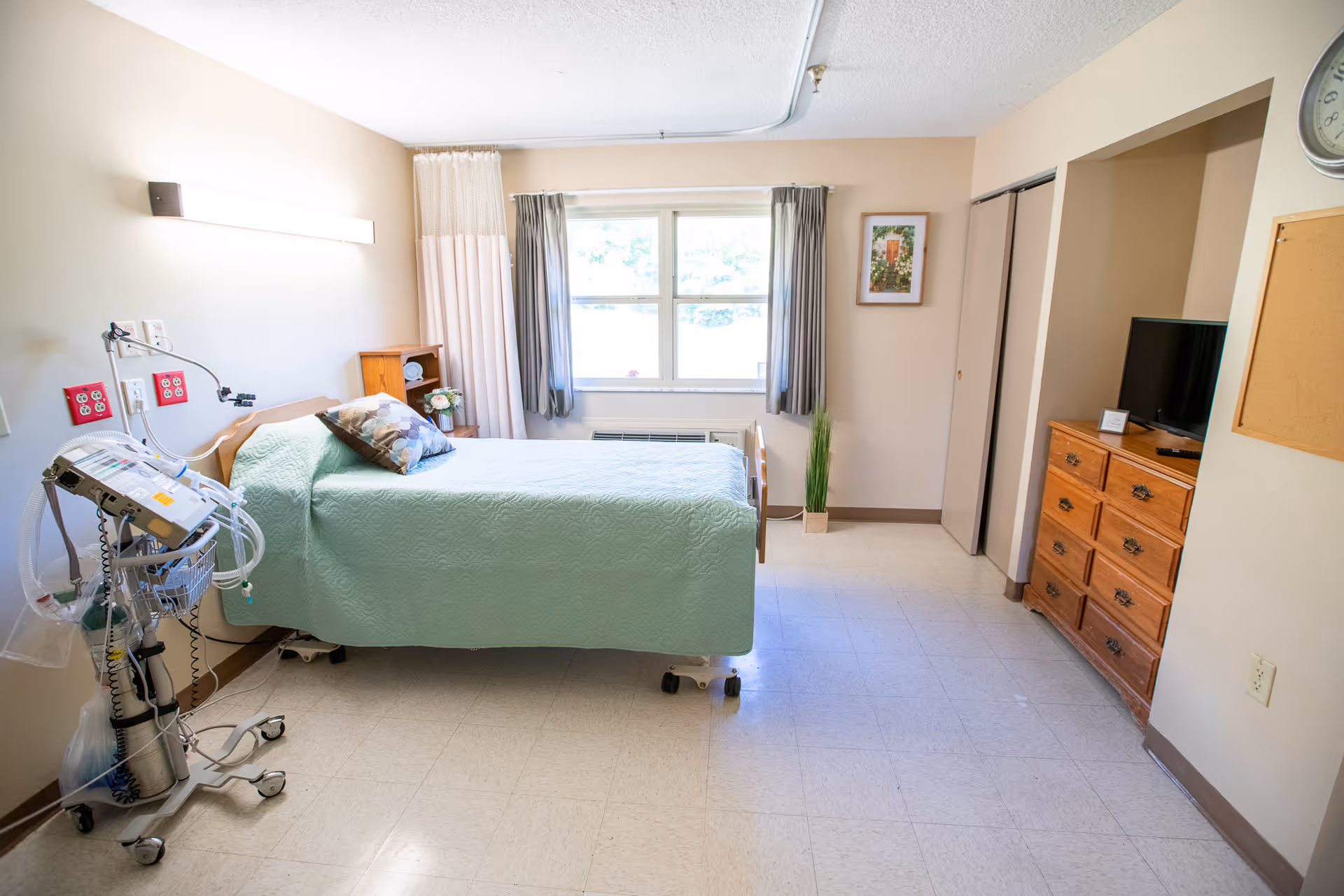 A clean and well-lit nursing facility bedroom with a hospital bed covered in a light green quilt, a pillow, and medical equipment on a stand next to the bed. The room has a window with curtains, a wooden dresser with a TV on top, a small plant, a framed picture on the wall, and a clock above a corkboard.