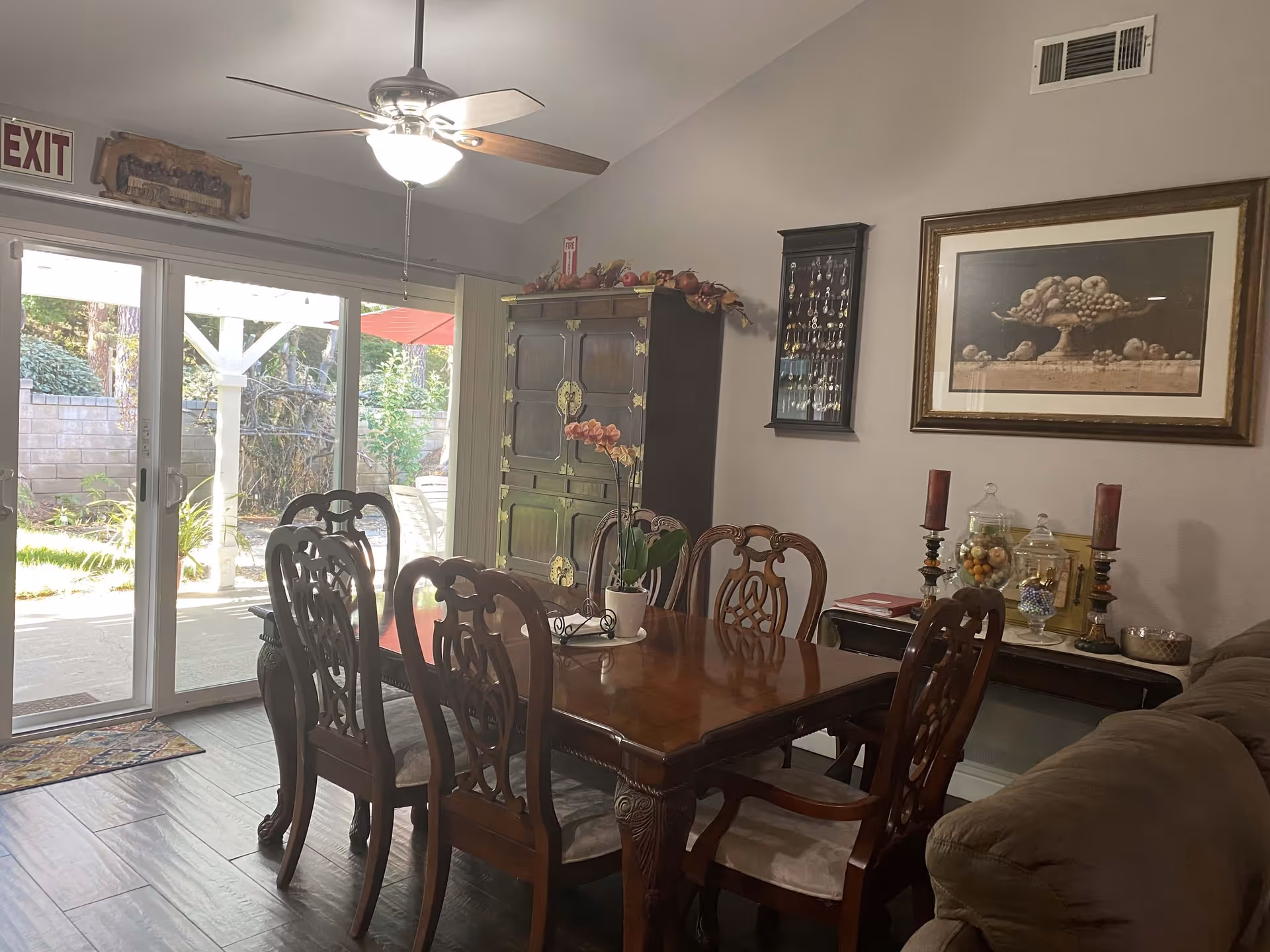 Dining room with a wooden table and six ornate chairs, a ceiling fan with light above, a large wooden cabinet decorated with artificial fruit, a glass sliding door leading to an outdoor patio, and a wall with framed artwork and decorative items including candles and glass jars.