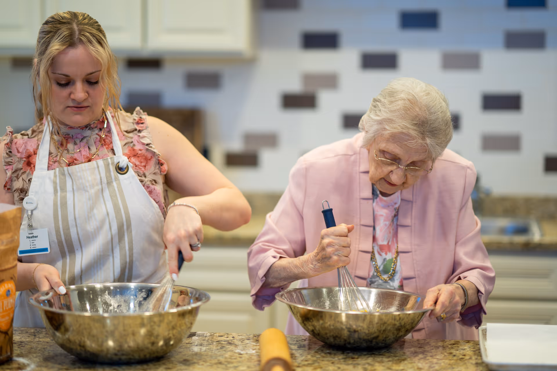 A younger woman and an elderly woman stand at a kitchen counter whisking ingredients in metal mixing bowls.