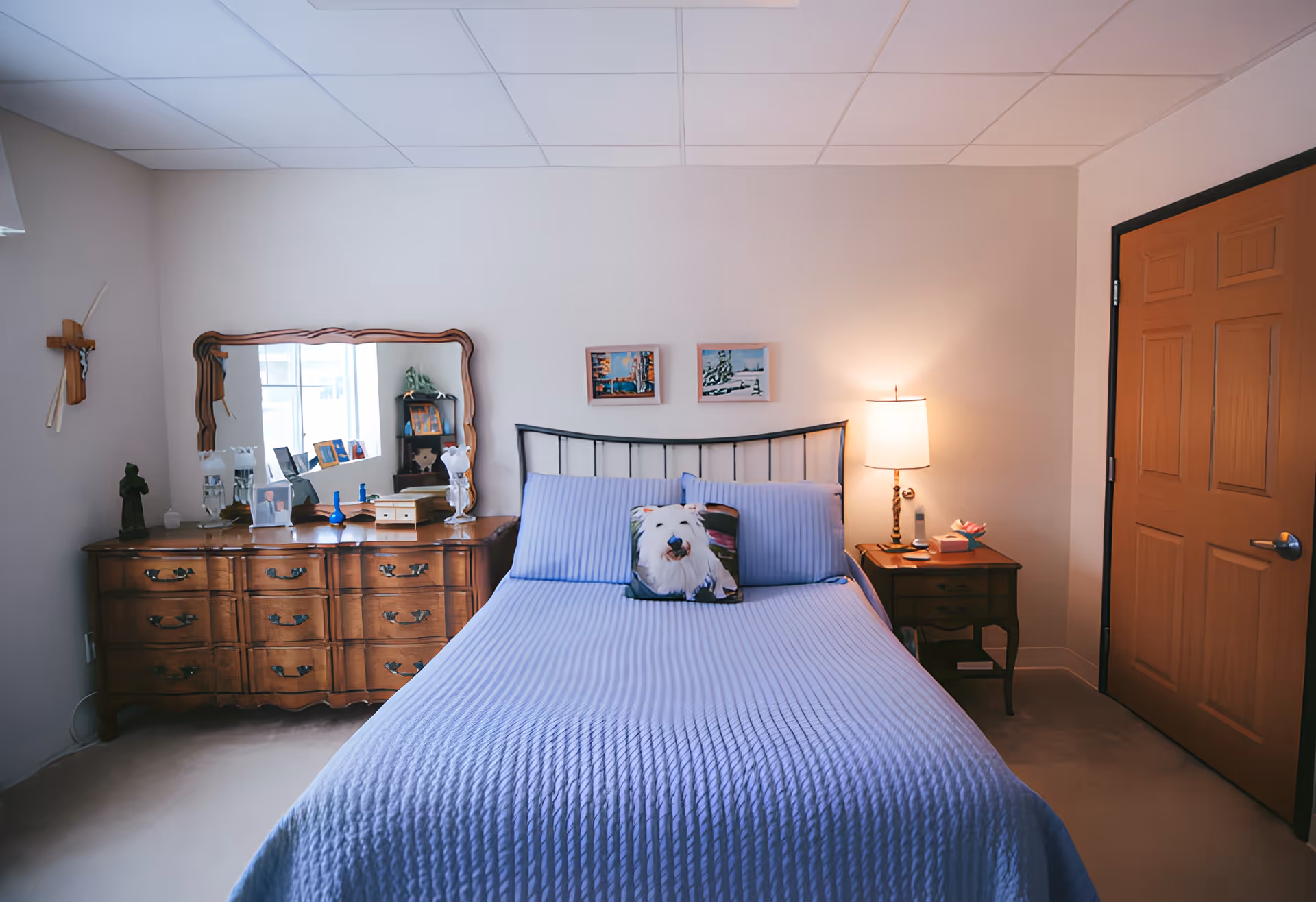 A cozy bedroom with a neatly made bed covered in a light blue quilt and matching pillows, including a decorative pillow with a dog image. To the left of the bed is a wooden dresser with a large mirror and various decorative items on top. On the right side of the bed is a wooden nightstand with a lamp, a tissue box, and other small items. The room has beige walls, a wooden door, and a crucifix hanging on the wall to the left.