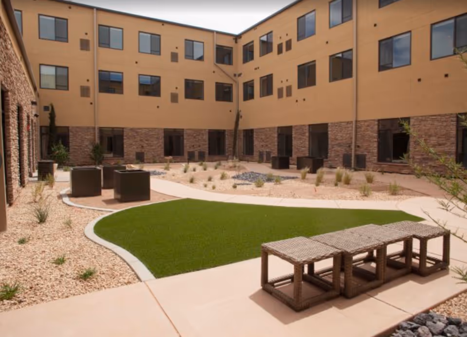 Sunny interior courtyard with gravel landscaping, a small artificial turf lawn, wicker benches and seating, surrounded by a three-story building.