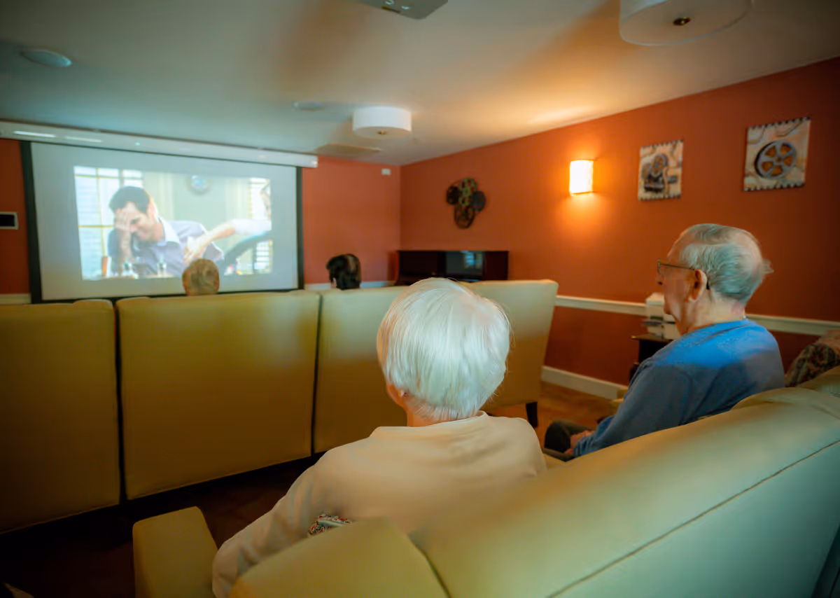 Elderly residents seated on sofas watching a movie projected on a screen in a communal lounge.