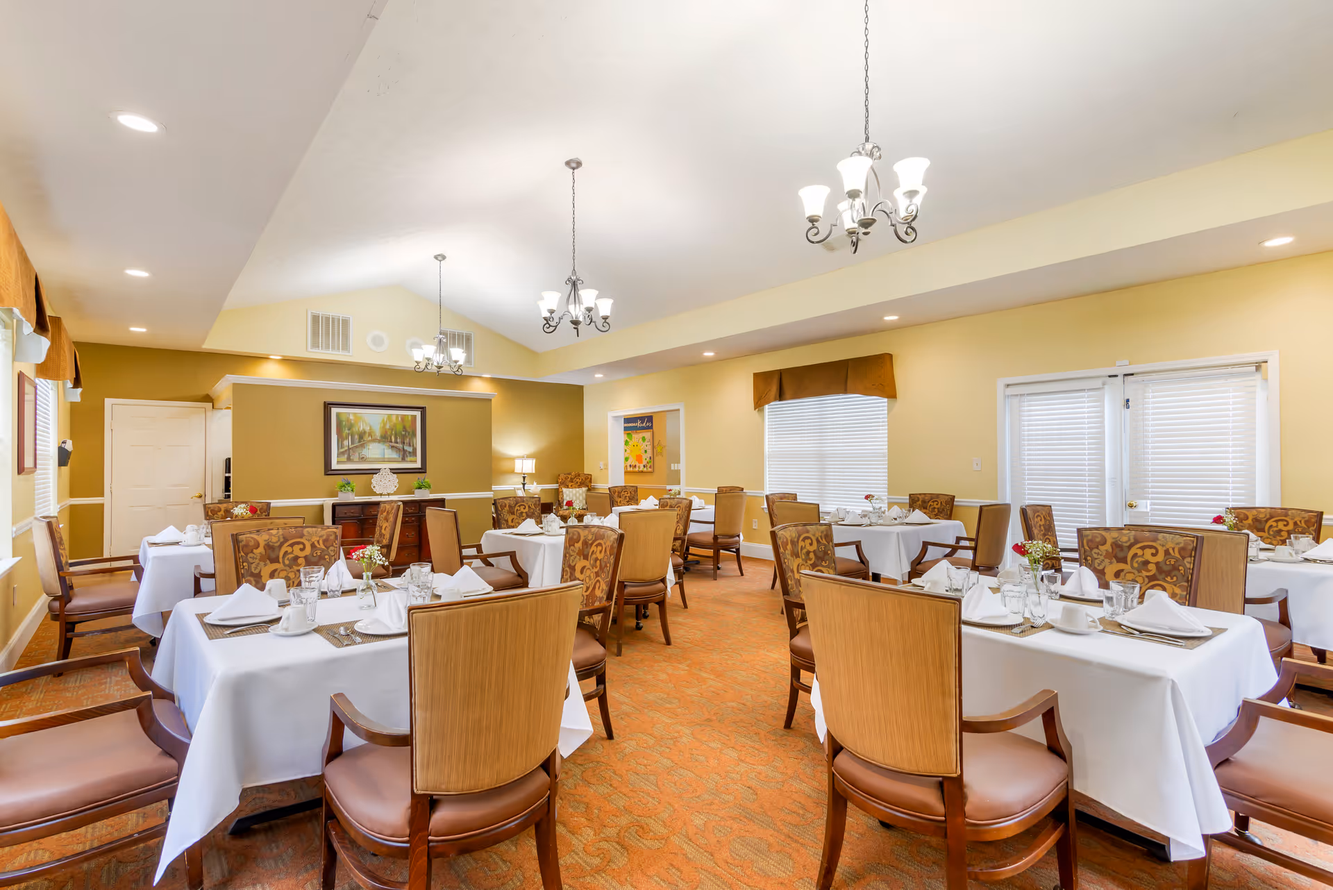 A well-lit dining room with multiple tables covered in white tablecloths, each set with napkins, cups, glasses, and small flower arrangements. The room features beige walls, patterned carpet, and several chandeliers hanging from a vaulted ceiling. There are large windows with blinds and valances, and a painting on one wall above a wooden sideboard.