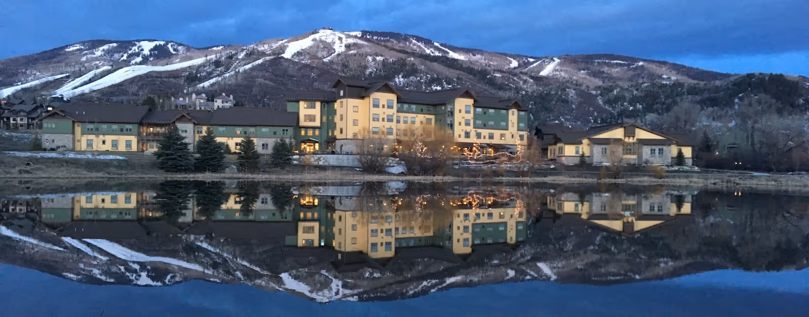 A large multi-story building with yellow and green exterior walls is situated near a calm body of water, reflecting the building and surrounding landscape. Behind the building, there are snow-covered mountains under a blue sky during dusk or dawn.