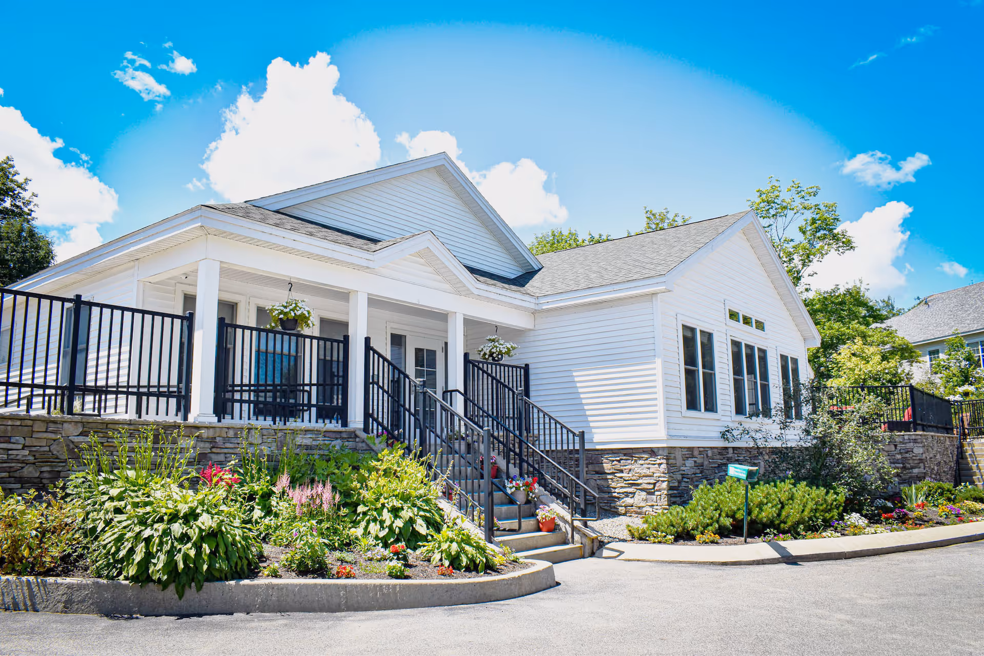 White single-story building with a covered porch, black railings, steps and landscaped flower beds under a bright blue sky.