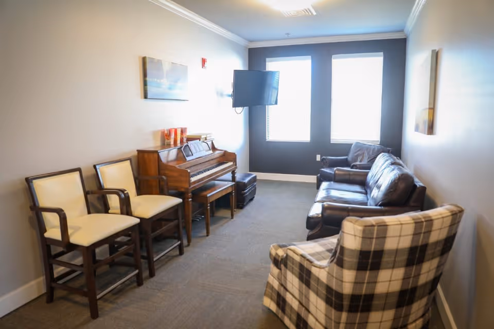 A cozy living room with a piano against one wall, two cream-colored chairs, a plaid armchair, and a dark leather sofa. Two large windows with blinds let in natural light, and a flat-screen TV is mounted on the wall between the windows. The walls are painted in neutral tones with a piece of artwork hanging above the piano.