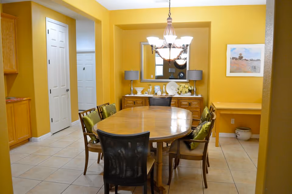 Dining room with an oval wooden table surrounded by chairs, yellow walls, a chandelier, and a sideboard with a mirror.
