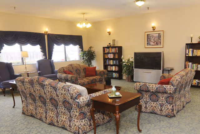 A cozy living room with patterned sofas and armchairs arranged around a wooden coffee table. The room has carpeted flooring, bookshelves filled with books, a large flat-screen TV, and windows with dark curtains letting in natural light. There are also potted plants and wall-mounted lights.