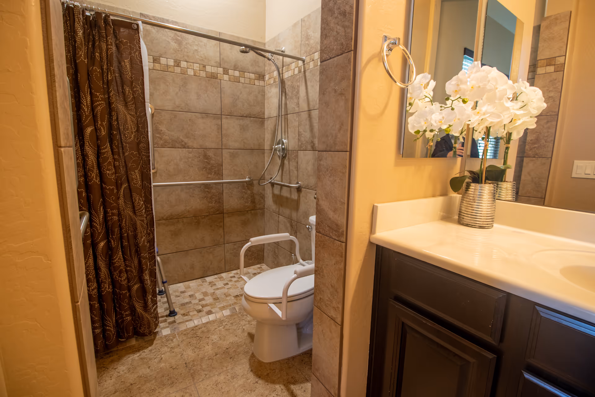 A bathroom with beige tiled walls and floor, featuring a walk-in shower with a brown patterned shower curtain. There are safety grab bars inside the shower and a toilet with white support rails. A white countertop with a dark wood cabinet below holds a silver vase with white flowers. A mirror is mounted on the wall above the countertop.