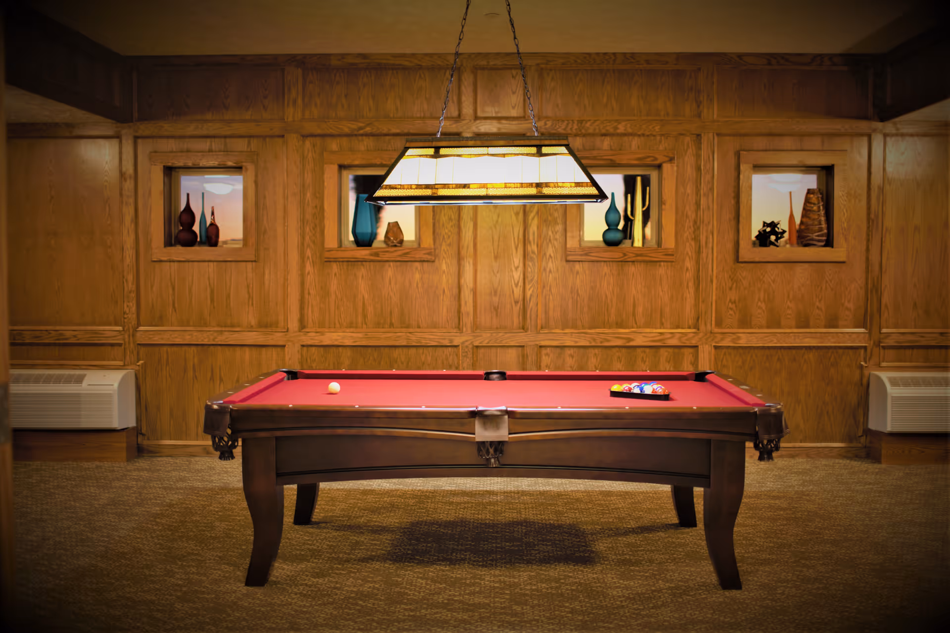 A pool table with a red felt surface in a room with wood-paneled walls. The pool table has a single white cue ball and a rack of colored balls set up. Above the table hangs a rectangular light fixture. The walls have three small recessed display windows with decorative vases and objects inside. There are two air conditioning units on either side of the room near the floor.