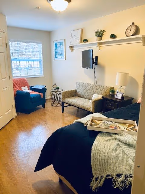 A cozy, well-lit room with a bed in the foreground covered with a black blanket and a light green knitted throw. There is a small beige loveseat against the wall with a mounted flat-screen TV above it. A dark wooden nightstand with a lamp, a framed photo, and a small plant is next to the loveseat. A blue armchair with a coral throw blanket is positioned near a window with white blinds. The walls are light-colored, and there is a shelf above the loveseat with decorative items including a clock and small plants. The floor is wooden.