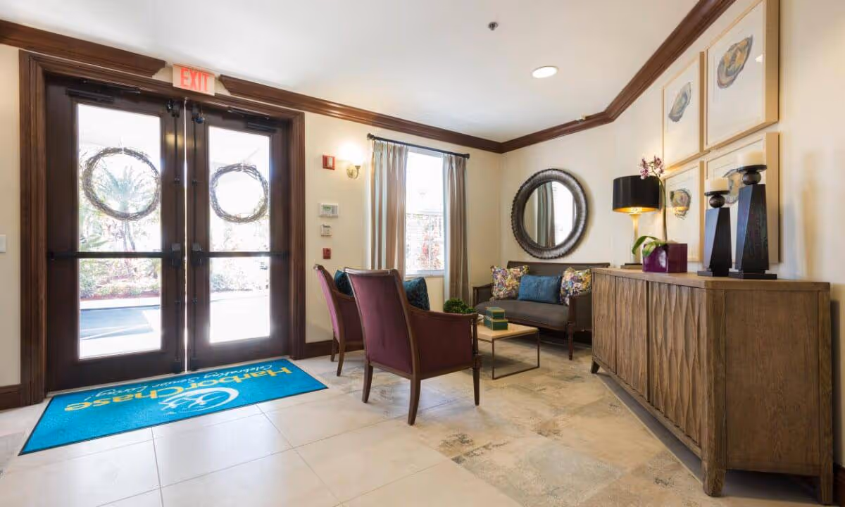 A bright and welcoming lobby area with double glass doors featuring wreath decorations. Inside, there are two purple armchairs and a gray sofa with colorful cushions arranged around a small coffee table. A wooden sideboard with decorative items, a lamp, and framed artwork is against the wall. A round mirror hangs above the sofa, and a blue floor mat with the HarborChase logo is placed near the entrance.
