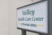 A white sign mounted on a post in front of a light-colored brick wall displaying the text 'Valley Health Care Center' and a phone number 276-646-8911.