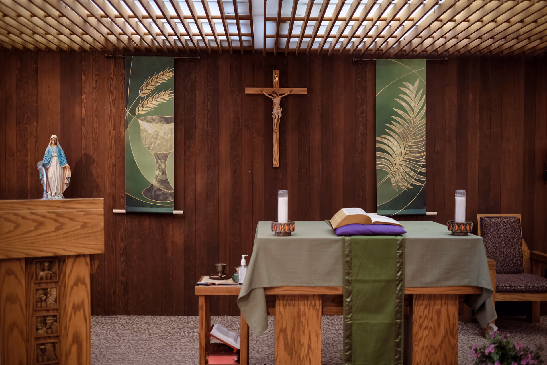 Interior of a small chapel with wooden walls and ceiling. A wooden altar is covered with a green cloth and holds an open Bible, two white candles, and a purple cloth. Behind the altar, a wooden crucifix is mounted on the wall, flanked by two green banners with religious symbols. To the left, there is a wooden lectern with a statue of the Virgin Mary. A chair is visible to the right of the altar.