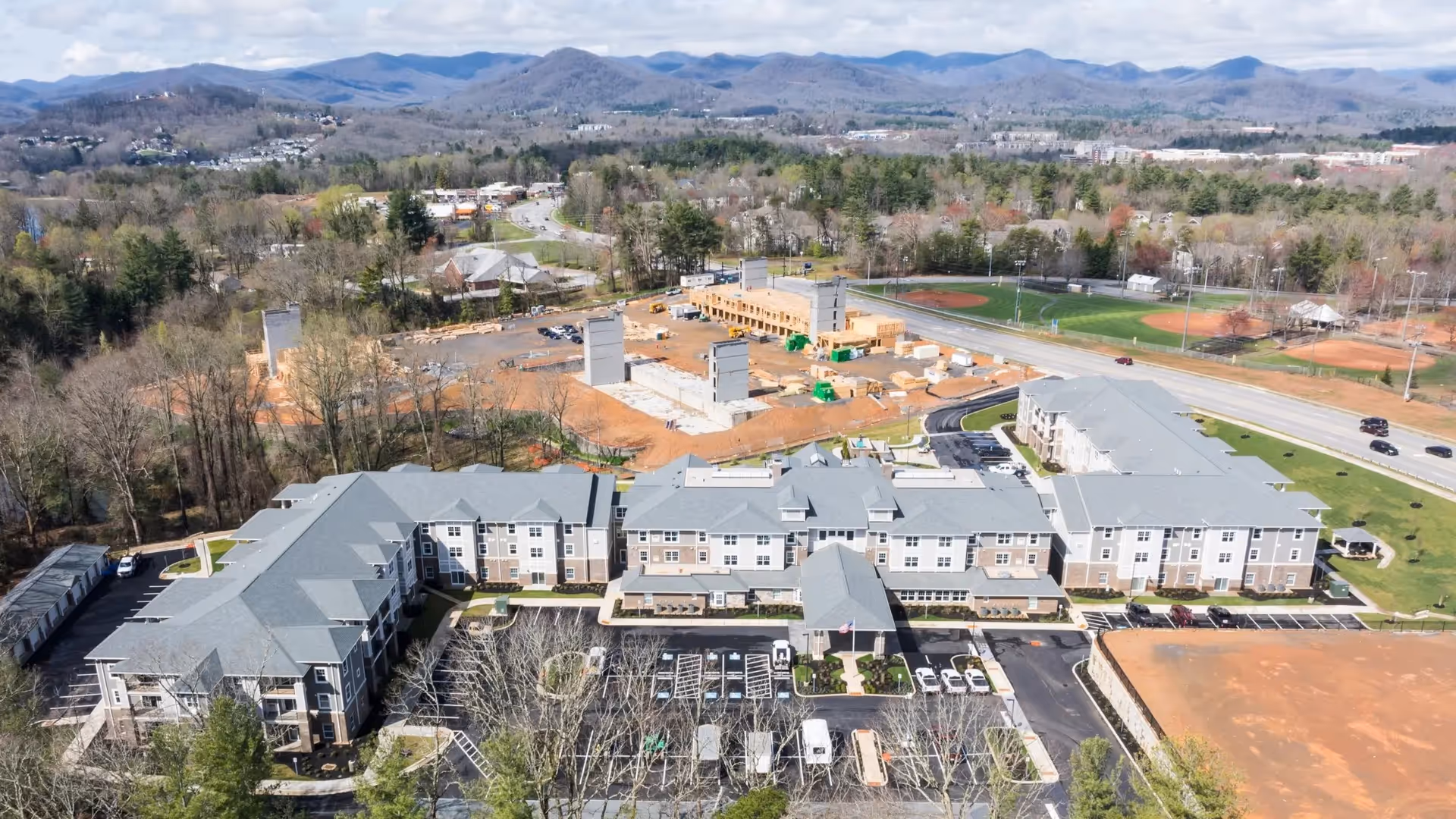 Aerial view of Pinnacle Springs Retirement Community showing a large, multi-wing building with gray roofs surrounded by parking lots, trees, and nearby construction. In the background, there are mountains and a few baseball fields adjacent to a road.
