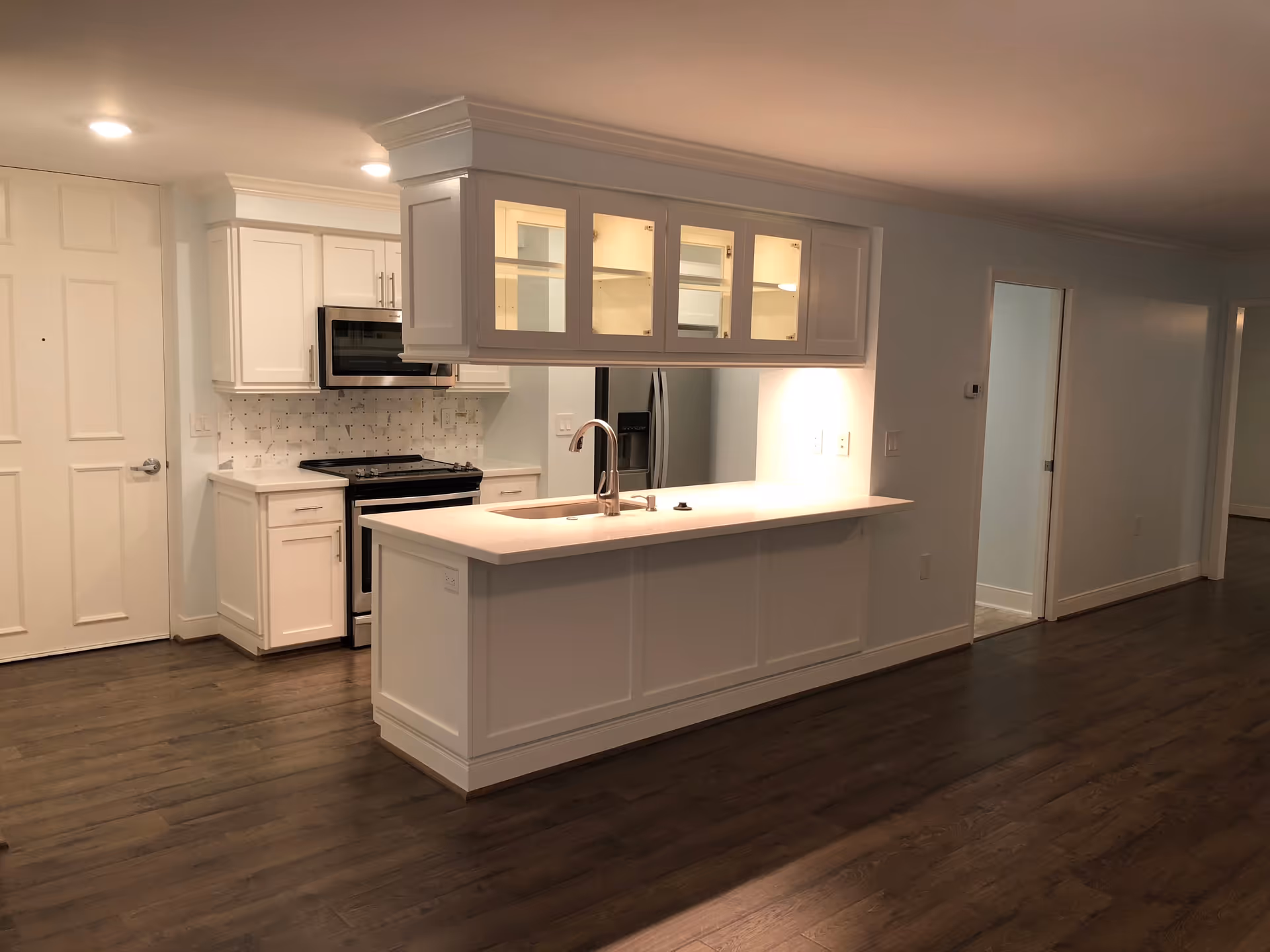 Interior view of a modern kitchen with white cabinetry, a kitchen island with a sink and faucet, stainless steel appliances including a microwave and refrigerator, and dark wood flooring. The kitchen is well-lit with recessed ceiling lights and has an open layout leading to other rooms.