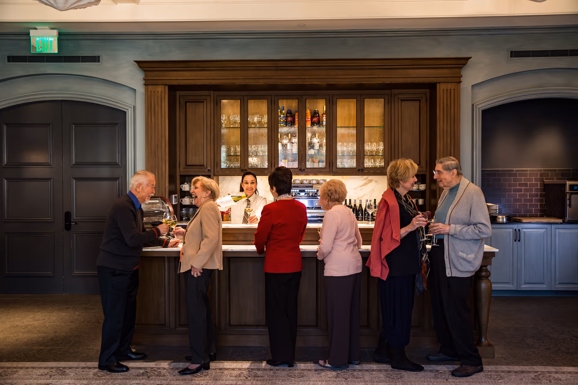 A group of six elderly people standing at a wooden bar counter inside a facility, engaging in conversation and holding drinks. Behind the counter, a bartender is smiling and serving drinks. The background features wooden cabinets with glass doors displaying bottles and glassware, and the setting has a warm, inviting atmosphere.